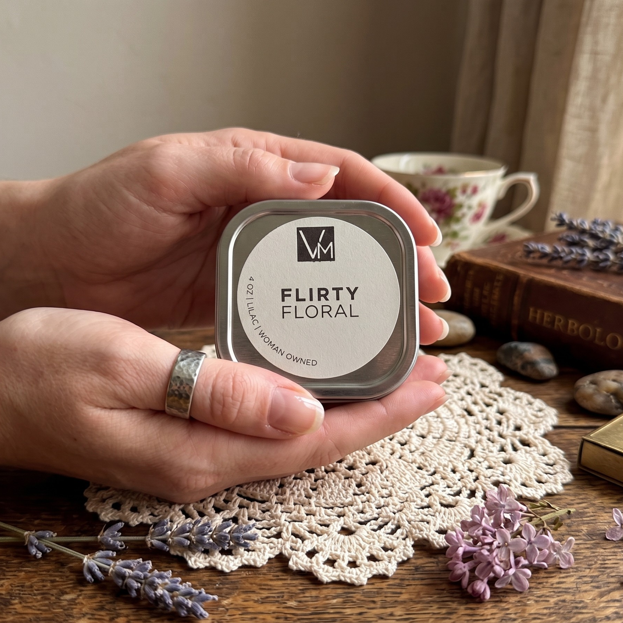 Hand holding a 'Flirty Floral' product container on a wooden surface with flowers and a teacup in the background.